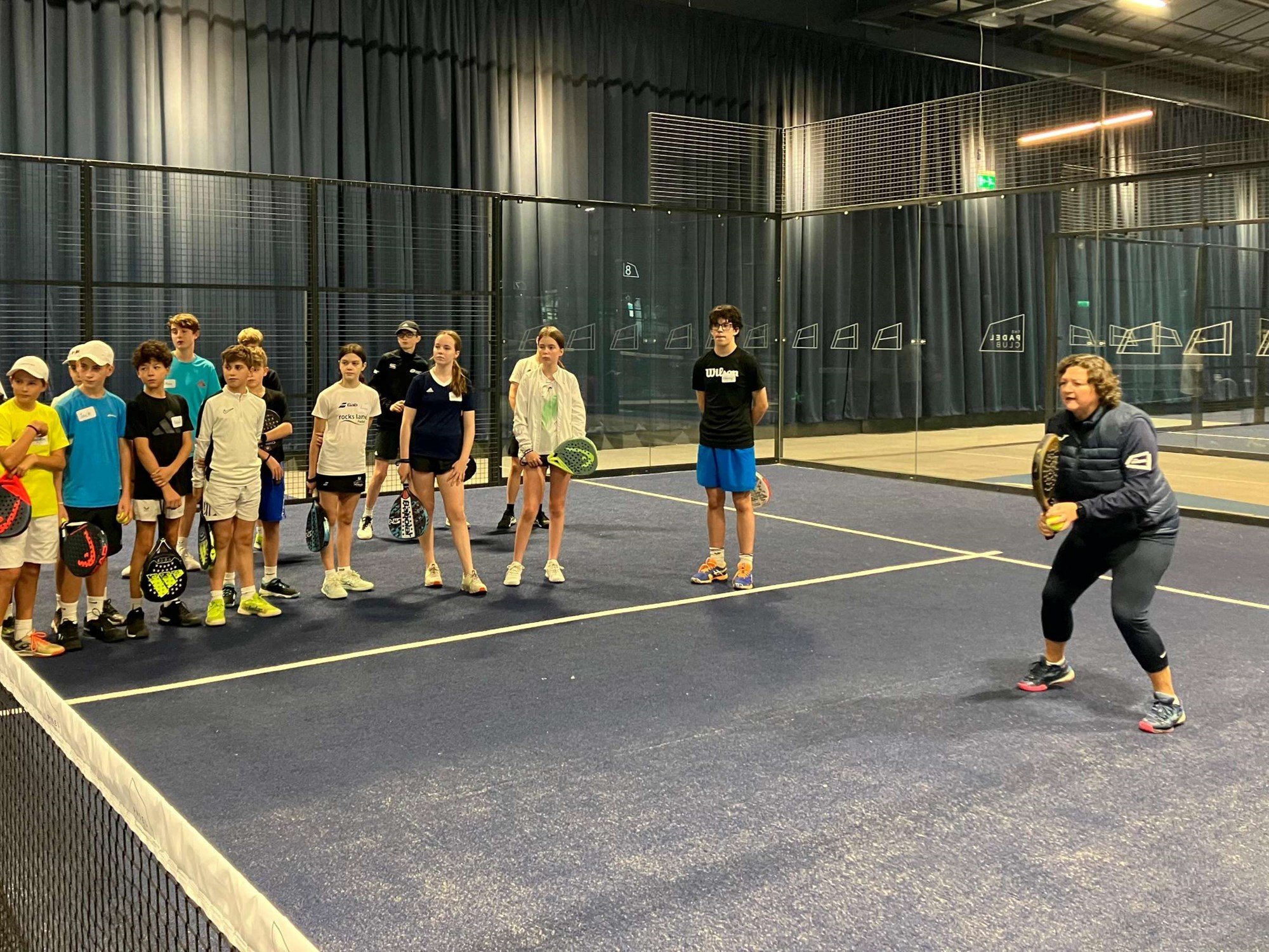 Coach Libby Fletcher crouching down preparing to return a shot while junior padel players watch on