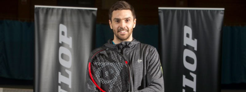 colin fleming  smiling for a picture  wearing all back with a racket in hand and dunlop  banners at the back