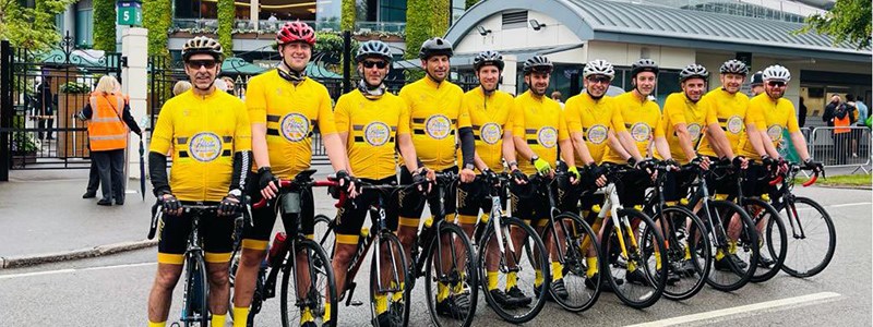 rutherglen peloton team standing on their bikes lined up outside wimbledon with yellow shirts and bike helmets on