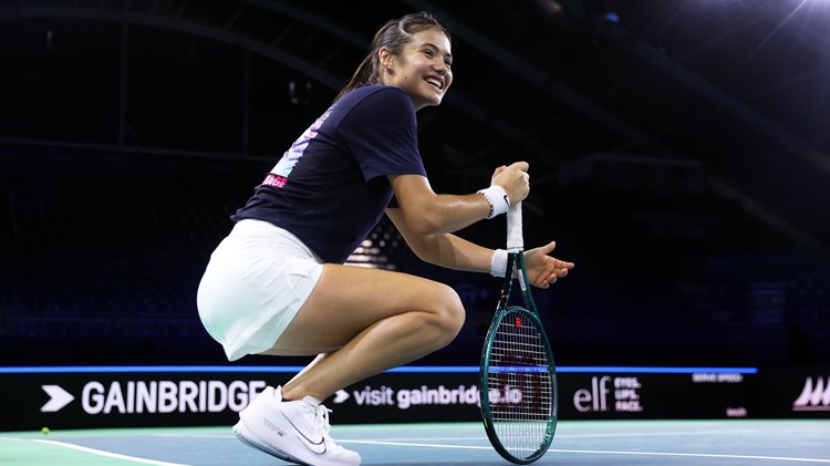 Emma Raducanu crouching down on court while smiling and holding her racket at the Billie Jean King Cup Finals in Malaga