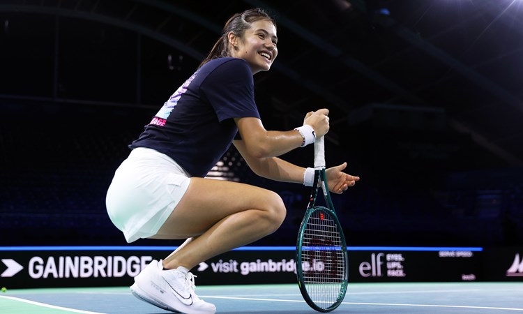 Emma Raducanu crouching down on court while smiling and holding her racket at the Billie Jean King Cup Finals in Malaga