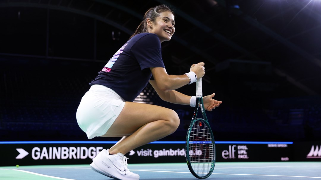 Emma Raducanu crouching down on court while smiling and holding her racket at the Billie Jean King Cup Finals in Malaga