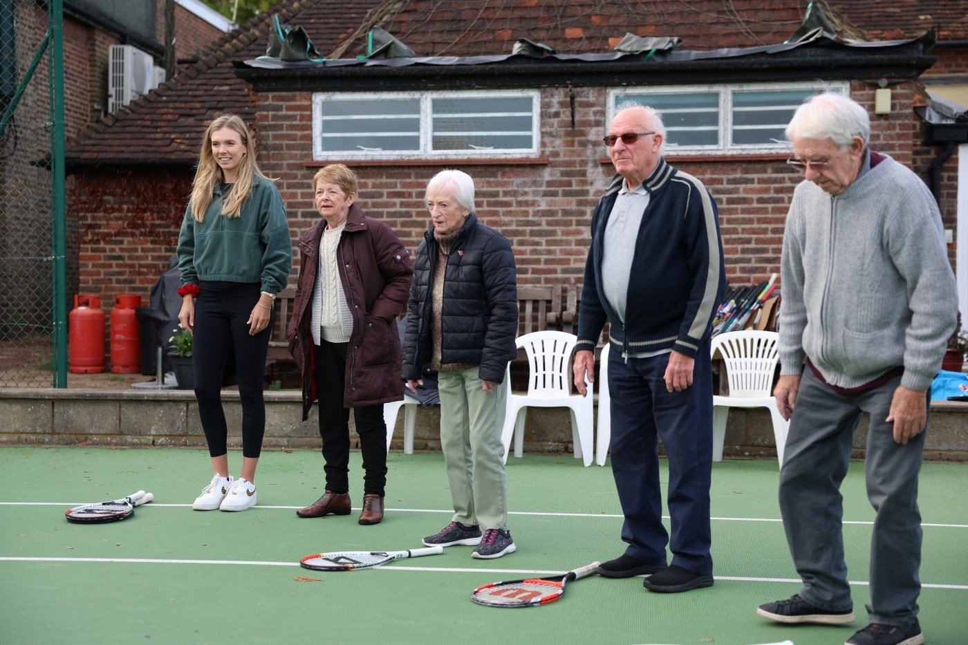 A group of five people, four elderly and one young person on the far left of the group, stand on a tennis court, with tennis rackets lying on the floor, in front of a brick building  