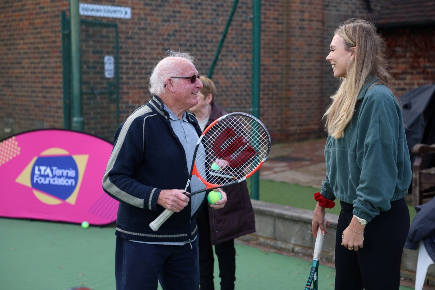 An elderly man holding a tennis racket and tennis ball talks to a younger owman stood next to him. Both have a smile on their face. A bright pink banner with the words 'LTA Tennis Foundation' is situated in he background