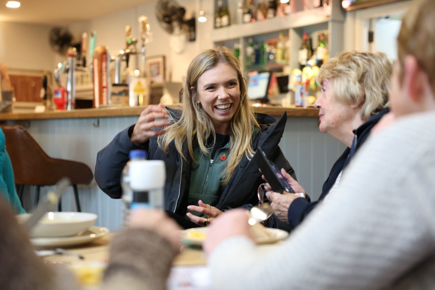 A woman is sat at a table talking with two enderly women on her left hand side, a smile on her face, with her right hand raised slightly in the air. She is wearing a black puffer jacket, unzipped, displaying a green jumper on which she is wearing a small red poppy badge.