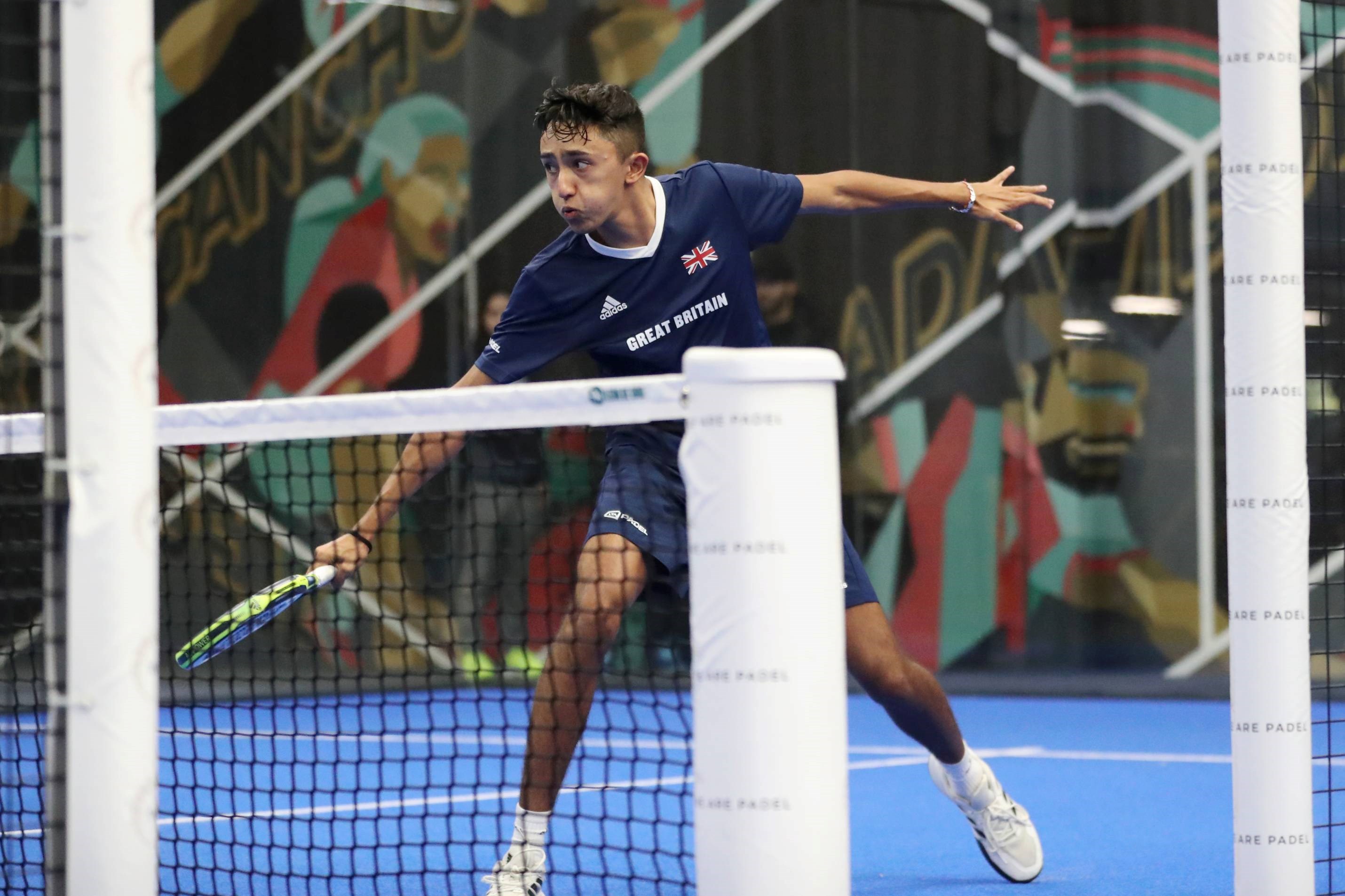 British padel player Nikhil Mohindra wearing a blue adidas top with a great Britain flag while hitting a backhand on a padel court