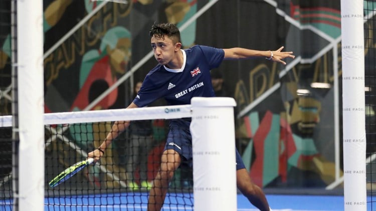 British padel player Nikhil Mohindra wearing a blue adidas top with a great Britain flag while hitting a backhand on a padel court