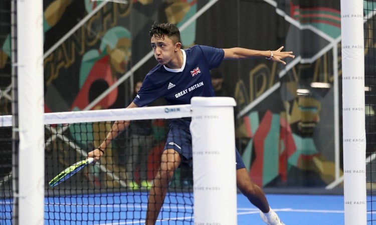 British padel player Nikhil Mohindra wearing a blue adidas top with a great Britain flag while hitting a backhand on a padel court