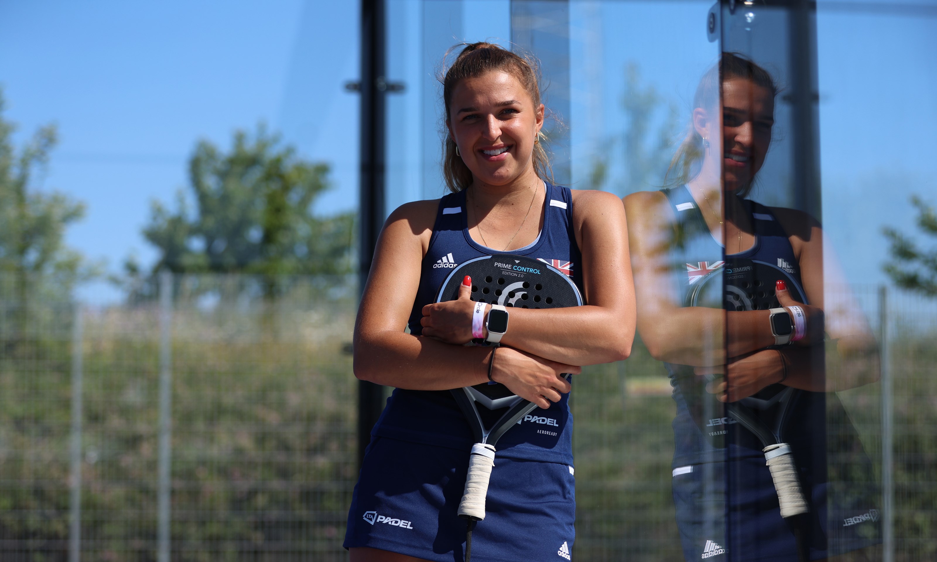 British padel player Catherine Rose leaning against the glass wall of a padel court wile holding her padel racket in her hands and wearing a blue 'LTA Padel'  kit