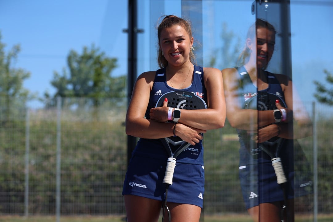 British padel player Catherine Rose leaning against the glass wall of a padel court wile holding her padel racket in her hands and wearing a blue 'LTA Padel'  kit