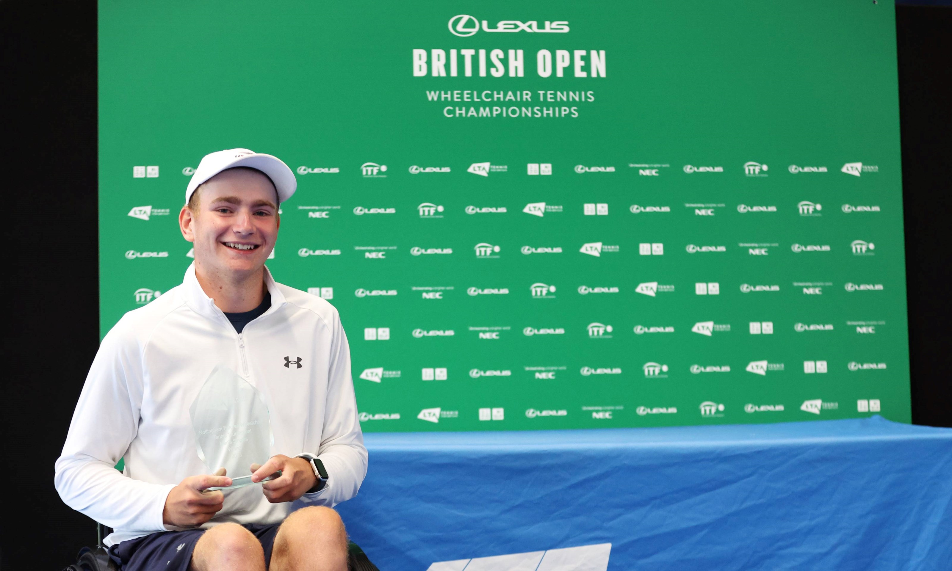 British wheelchair tennis player Andrew Penney smiling while sat in his wheelchair in front of a 'Lexus British Open Wheelchair Tennis Championships' branding board