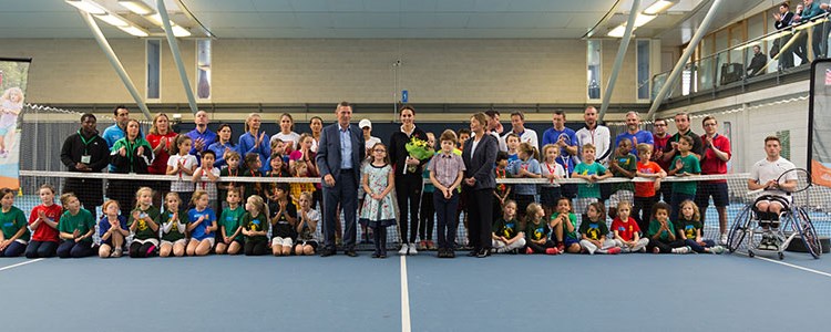 Duchess of Cambridge standing in the middle of a group of kids and coaches on a tennis court by the net