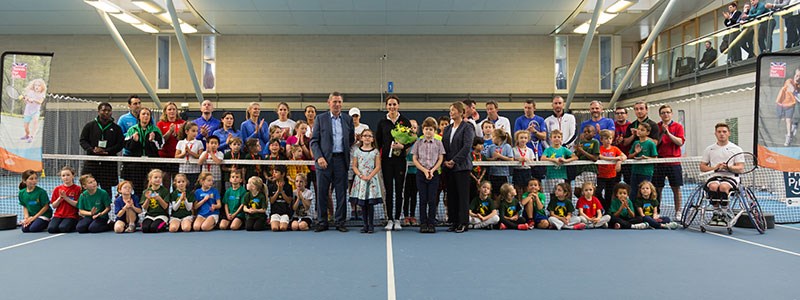 Duchess of Cambridge standing in the middle of a group of kids and coaches on a tennis court by the net