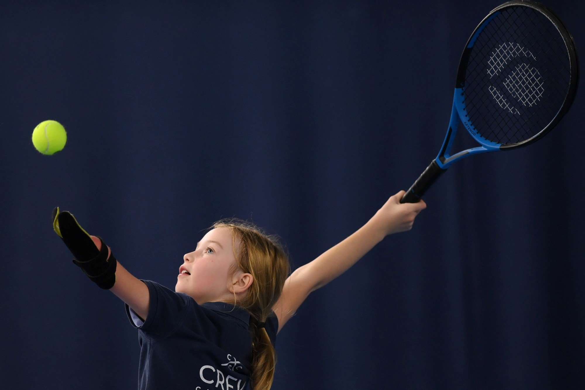 A picture of a young girl with an upper limb impairment raising her hand to serve on court