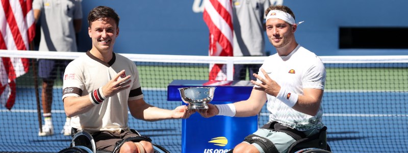 hewett and reid holding trophy in one hand and  other hand holding up four fingers  with us open logo at the back