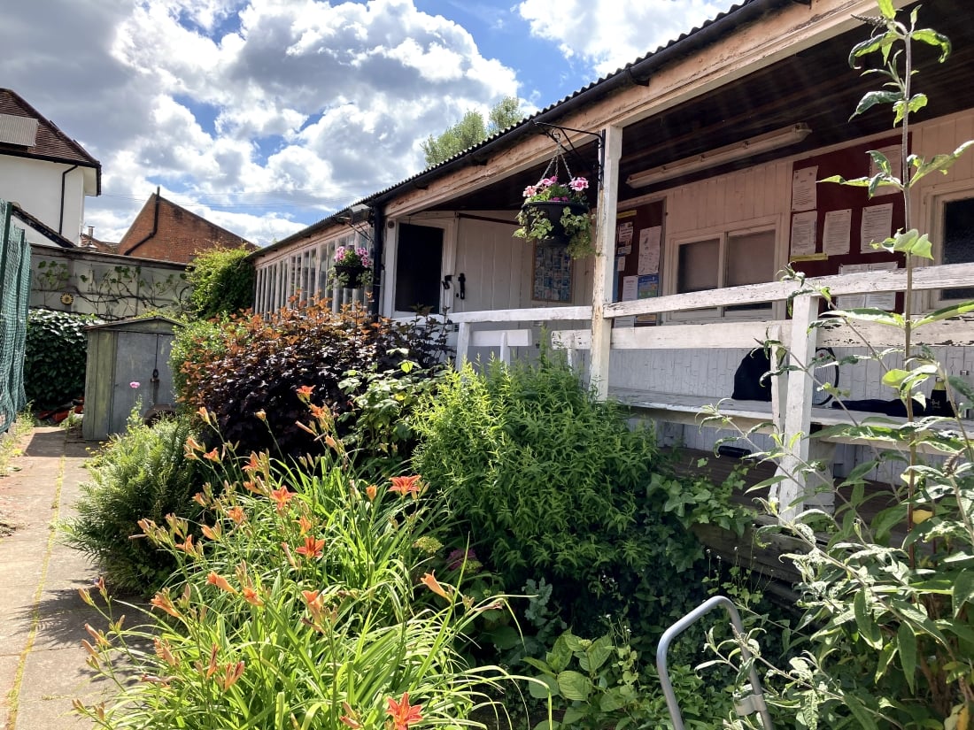 A large amount of flowers and green foliage is planted in front of a wooden building painted white. A path leads away down the left hand side of the plant bed