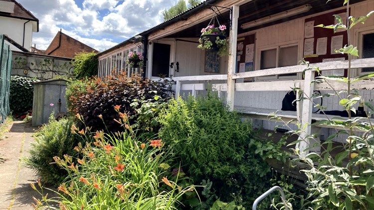 A large amount of flowers and green foliage is planted in front of a wooden building painted white. A path leads away down the left hand side of the plant bed