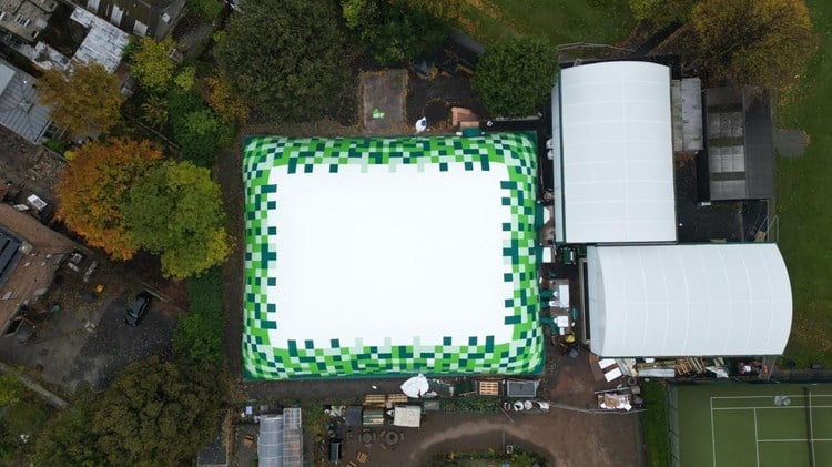 Aerial view of a rectangular dome whose edges are green in colour. To the right hand side there are two domes and part of a tennis court. Trees surround the dome.