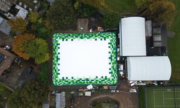 Aerial view of a rectangular dome whose edges are green in colour. To the right hand side there are two domes and part of a tennis court. Trees surround the dome.