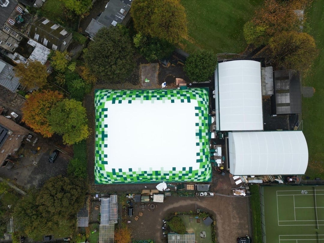 Aerial view of a rectangular dome whose edges are green in colour. To the right hand side there are two domes and part of a tennis court. Trees surround the dome.